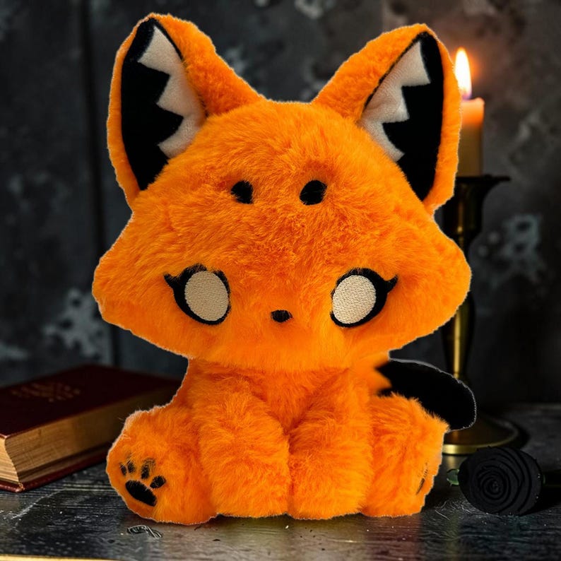 May include: An orange plush fox with black accents, including ear tips, paws, and a tail. The fox has large, round eyes and a friendly expression. The plush toy is sitting upright, with a paw print on one foot. A candle and a book are in the background.