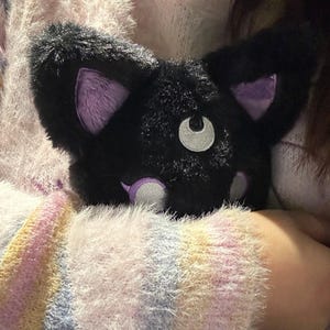 May include: A plush black cat-shaped pillow with purple inner ears and accents. The cat has a white crescent moon-shaped eye and a small white nose. The pillow is soft and fluffy, perfect for cuddling.