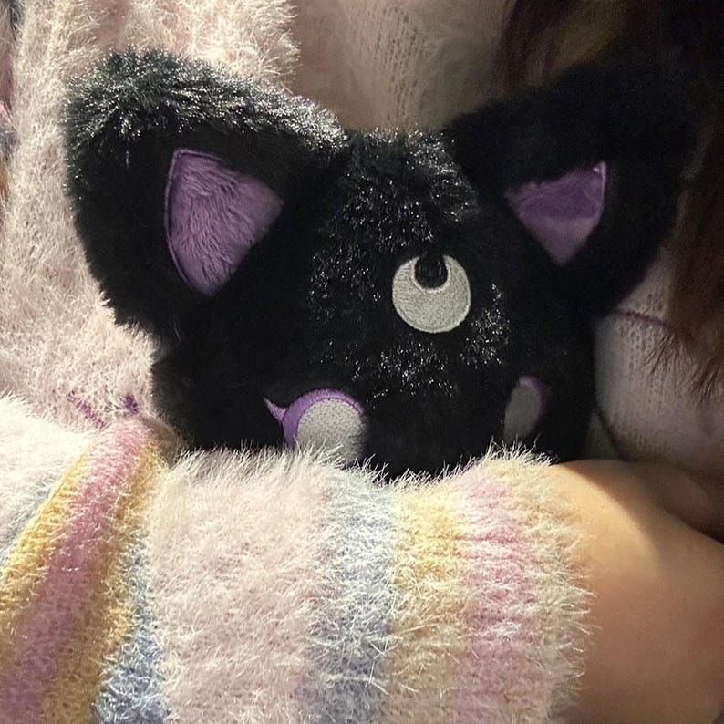 May include: A plush black cat-shaped pillow with purple inner ears and accents. The cat has a white crescent moon-shaped eye and a small white nose. The pillow is soft and fluffy, perfect for cuddling.