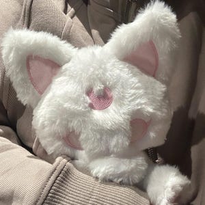 May include: A fluffy white plushie shaped like a cat's head with pink inner ears and facial details. The plushie has a zipper and is made of soft, textured material. The cat plushie is a cute accessory.