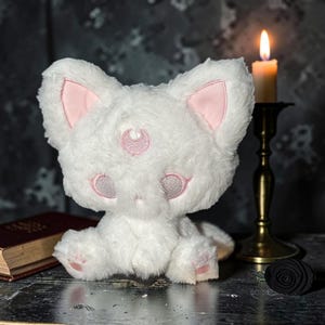 May include: A fluffy white cat plushie with pink inner ears and facial details. It features a crescent moon emblem on its forehead. A lit candle in a brass holder and a book are in the background.