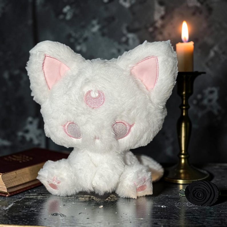 May include: A white plush cat with pink accents on the ears, eyes, and forehead. The cat has a crescent moon design on its forehead. A lit candle in a brass holder and a book are in the background.