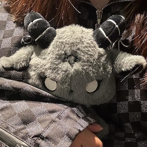 May include: A gray plush toy shaped like a horned creature with large white eyes and black horns with white stripes. The plush is soft and fluffy, with a detailed design. The toy is held against a black and gray checkered jacket.