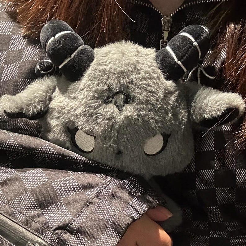 May include: A gray plush toy shaped like a horned creature with large white eyes and black horns with white stripes. The plush is soft and fluffy, with a detailed design. The toy is held against a black and gray checkered jacket.