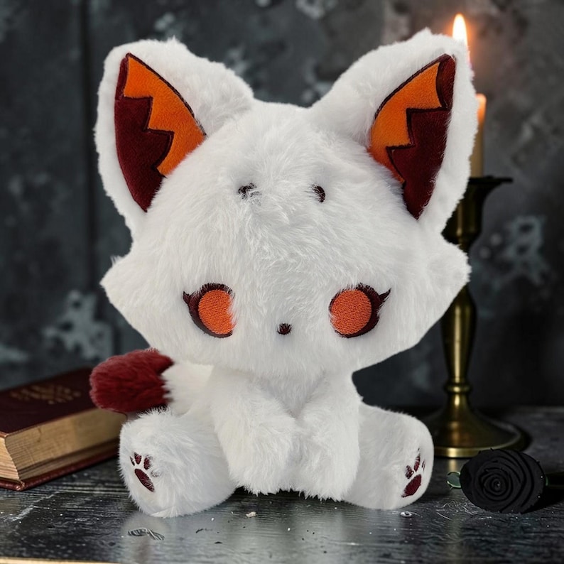 May include: A white plush fox with orange and burgundy accents. The fox has large, round orange eyes, matching ear details, and a burgundy tail. The plush toy is sitting with its paws forward. A small book and candle holder are in the background.