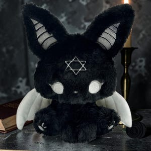 May include: A black plush bunny with white accents, large white eyes, and a six-pointed star on its forehead. The bunny has white wings and is posed on a dark surface. The bunny's ears have white stripes.