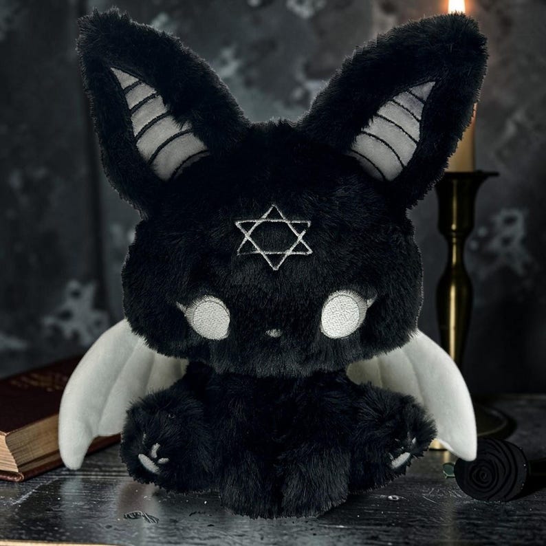 May include: A black plush bunny with white accents, large white eyes, and a six-pointed star on its forehead. The bunny has white wings and is posed on a dark surface. The bunny's ears have white stripes.