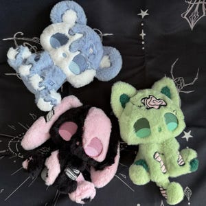 Pode incluir: Três brinquedos de pelúcia: um urso azul e branco, um coelho preto e rosa e um gato verde. O urso e o coelho têm desenhos de caveira. O gato tem um cérebro e detalhes de bandagem.