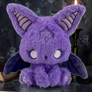 May include: A plush purple bat-like creature with large ears, wings, and a star emblem on its forehead. The toy has white eyes, a fluffy texture, and is sitting. The background includes a candle and a book.
