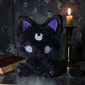 May include: A black plush cat with purple accents on the ears and paws, and light blue eyes. The cat has a third eye with a crescent moon design. A lit candle in a brass holder and a small book are in the background.