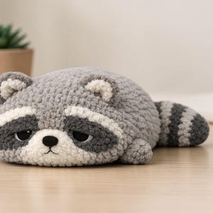 May include: A handmade, plush raccoon toy in shades of gray, white, and black. The raccoon is lying down with a striped tail and a sad expression. The toy is on a light-colored wooden surface.