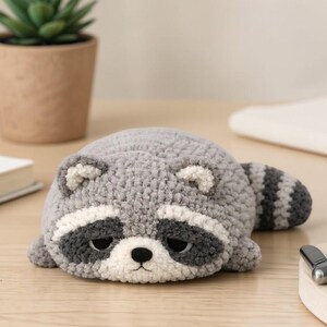 May include: A handmade, gray crochet raccoon plushie with black and white facial markings and a striped tail. The raccoon is lying down on a wooden surface. A small potted plant and a book are in the background.