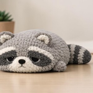 May include: A handmade, plush raccoon toy in shades of gray, white, and black. The raccoon is lying down with a sad expression, featuring a white face with black eye markings and a striped tail. The toy is on a light-colored wooden surface.
