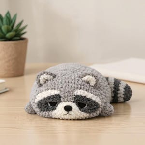 May include: A handmade crochet raccoon toy in gray, white, and black, lying on a wooden surface. The raccoon has a striped tail and a sad expression. A small potted plant and a book are in the background.