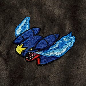 May include: An embroidered patch featuring a stylized dark blue creature with a yellow star-shaped marking on its head, a red mouth with visible white teeth, and flowing light blue flame-like accents extending from its body edges.