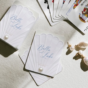 May include: A deck of playing cards and loose cards featuring a white shell-shaped embossed design, the text Bella and Luke, and the date 09.02.2026. Each card is accented with a small white pearl. Two gold-toned earrings with white pearls are included.