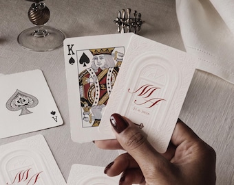 May include: A set of custom playing cards featuring a white card back with an embossed archway design, the red monogram MA, and the date 21.6.2026. One card shows the King of Spades, while another displays the Ace of Spades. The cards have a matte finish.
