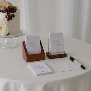 Puede incluir: Un pastel blanco decorado con flores blancas, peras y uvas sobre un soporte de cristal, junto a dos tarjeteros de madera con tarjetas que dicen Bella and Luke, una tarjeta suelta y un marcador negro sobre un mantel blanco.