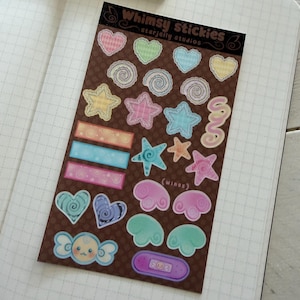 May include: A rectangular sticker sheet with a brown polka dot background featuring various pastel-colored designs including hearts, stars, swirls, rectangular labels, wings, and a character face. The header text reads Whimsy stickies starjelly studios.