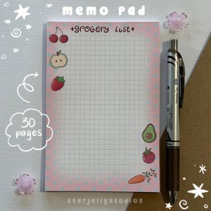 May include: A rectangular memo pad with a pink border featuring blue polka dots and a grid interior. The text "grocery list" is printed at the top. Illustrations of cherries, an apple, a strawberry, an avocado, a tomato, and a carrot are included.