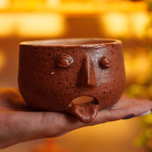 Könnte beinhalten: Eine kleine, runde Keramikschale in Terrakottafarbe mit einem skulptierten Gesicht, das zwei erhabene Augen, eine markante Nase und einen offenen Mund mit herausgestreckter Zunge aufweist. Die Oberfläche ist durchgehend gesprenkelt.