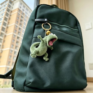 May include: A dark green backpack featuring a textured surface and a zippered front pocket. Attached to the zip pull is a gold-toned metal keyring with a small yellow bell and a plush, light green lizard figure with a wide-open mouth and pink tongue.