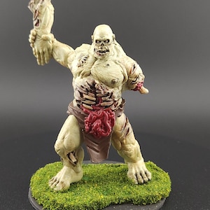 Op de afbeelding: Een handgeschilderde miniatuur zombiefiguur met zichtbare ribben en een losgemaakte arm. De figuur is geschilderd in tinten groen, bruin en rood en staat op een basis bedekt met groene flocking. De zombie staat in een dreigende houding.
