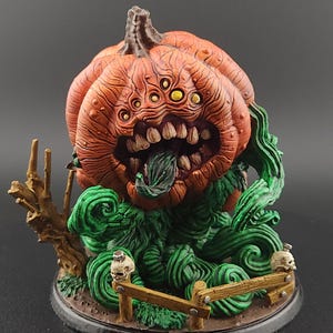May include: A detailed, orange pumpkin-like sculpture with multiple eyes and a gaping mouth, surrounded by green vines and a small wooden fence. Two small skulls are placed on the base. The sculpture has a dark base and is set against a dark background.
