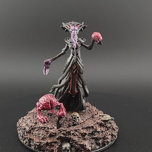 Op de afbeelding: Miniatuur fantasyfiguur van een donker gekleed wezen met een kroonachtig hoofd, dat een hersen vasthoudt. Het figuur staat op een getextureerde basis met schedels en een kleiner wezen. Het kleurenpalet omvat zwart, paars en rood. De figuur is ongeveer 10 cm hoog.