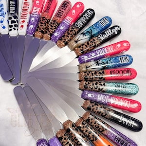 May include: A collection of nail swatch sticks with various designs and names. The nail tips feature glitter, animal prints, and tropical scenes. Names like "Kassidy", "Brittany", and "Devin" are written on the nail tips. The sticks have a silver metal handle.