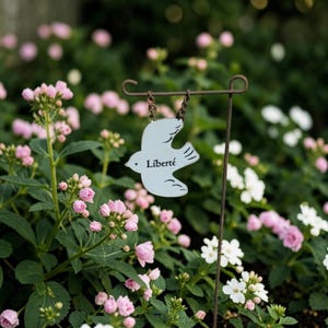 May include: A light blue metal bird ornament hanging from a thin brown metal garden stake by two small chains. The bird shape features the word Liberté printed in black on its center, with stylized wing and tail feather details etched into the surface.