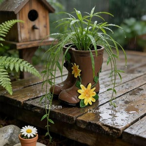 May include: Brown ceramic boot-shaped planter with yellow flower accents, holding a green plant. The planter is 11.5 cm tall and 12 cm wide. A small potted daisy sits nearby. The scene is set outdoors on a wooden surface.