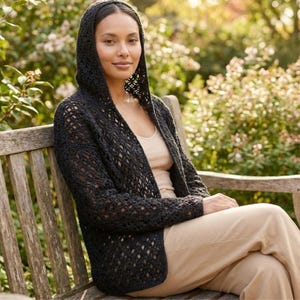 May include: A black open-weave hooded cardigan. The cardigan is worn over a light-colored top and tan pants. The person is sitting on a wooden bench in a garden setting with soft sunlight.