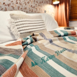 May include: A striped blanket in shades of teal, orange, brown, and white is draped over a bed. The blanket has a personalized embroidered name and tree design. A decorative pillow with fringe and a striped pattern is also on the bed.