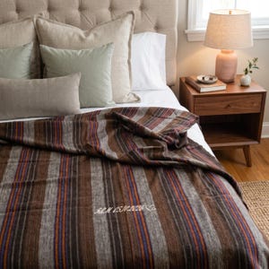 May include: A woven throw blanket featuring vertical stripes in shades of brown, gray, orange, and blue. The fabric is embroidered with the white text "S.L.M 05.10.2026" followed by a small heart outline, positioned near the center of the blanket.