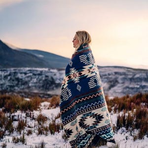 May include: A woven blanket featuring a dark blue base with a repeating geometric pattern in cream, brown, and teal. The design includes diamond shapes, horizontal stripes, and zigzag lines across the fabric, which is draped over the shoulders.