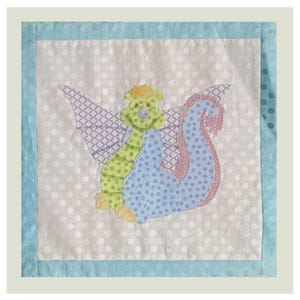 May include: A square fabric quilt block featuring a central applique of a mythical dragon creature. The dragon has a green polka-dot body, blue polka-dot tail, and purple patterned wings. The background is white with light gray polka dots.