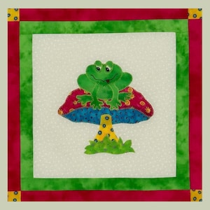 May include: A square fabric quilt featuring a green frog sitting on a colorful mushroom. The mushroom has a red top with yellow and blue accents. The quilt has a white background with a green, red, and yellow border.