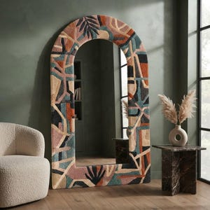 May include: A tall, arched floor mirror featuring a thick, textured frame covered in a tufted, multi-coloured fabric. The frame displays an abstract geometric pattern in shades of rust, teal, cream, black, and dusty pink, including leaf motifs.