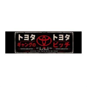 Puede incluir: Una pegatina rectangular negra con un logotipo rojo de Toyota, texto en japonés y el texto en inglés Toyota Gang Bitch Est. 2024. Debajo, la frase Choose the path with no road está impresa en blanco, enmarcada por un borde fino y desgastado.