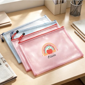 May include: Three rectangular, semi-transparent mesh zip pouches in pink, blue, and white. The pink pouch features a graphic of a rainbow, a pencil, and a red apple with the name Olivia printed in black script below the design.