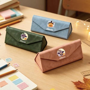 May include: Three triangular eyeglass cases in blue, green, and pink. Each case features a Halloween-themed sticker with a bat or pumpkin and the name Olivia printed in white. The cases have a smooth, matte finish and a magnetic flap closure.