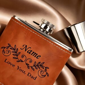 Może przedstawiać: Brązowy skórzany flakon z srebrną nakrętką i małym srebrnym kubkiem. Na flakonie wygrawerowano słowo "Name" i frazę "Love You, Dad" z dekoracyjnym kwiatowym wzorem.