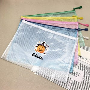 May include: A stack of five rectangular mesh zipper pouches in light blue, dark blue, pink, yellow, and green. The top light blue pouch features a graphic of a jack-o'-lantern wearing a witch hat with the name Olivia printed in black text below.