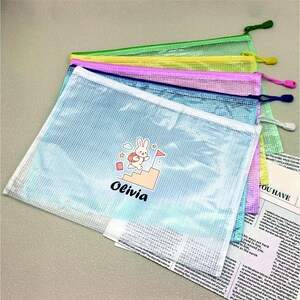 May include: A stack of four rectangular mesh zipper pouches in light blue, blue, pink, and yellow. The top light blue pouch features a graphic of a cartoon rabbit climbing stairs with the name Olivia printed in black text below the illustration.