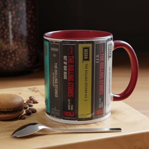 May include: A ceramic mug featuring a design of The Rolling Stones album covers. The mug has a burgundy interior and handle. A silver spoon and a macaron are on a wooden surface next to the mug.