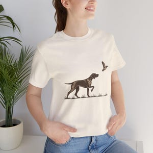 Peut inclure: Un t-shirt à manches courtes de couleur crème arborant une illustration graphique marron foncé représentant un chien d'arrêt debout dans l'herbe, une patte levée, regardant un petit oiseau volant dans les airs au-dessus de sa tête.