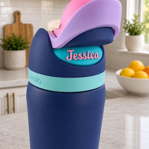 May include: A dark blue water bottle with a light blue band and a multi-colored lid featuring pink and lavender accents. The name Jessica is printed in pink letters on a teal oval plaque on the front. The brand name Owala is visible in light blue.