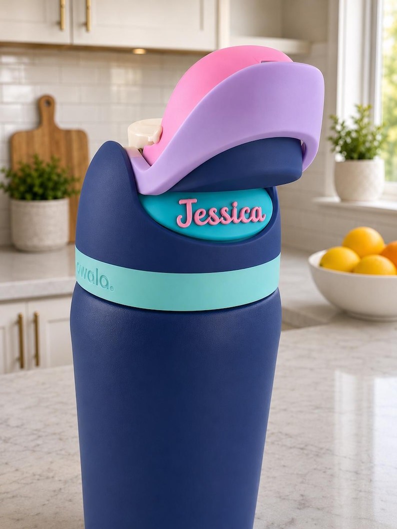 May include: A dark blue water bottle with a light blue band and a multi-colored lid featuring pink and lavender accents. The name Jessica is printed in pink letters on a teal oval plaque on the front. The brand name Owala is visible in light blue.
