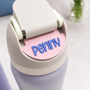 May include: A light purple water bottle with a beige lid featuring a hinged flip-top mechanism. A semi-circular pink name tag is attached to the front of the lid, displaying the name penny in raised, bold, dark blue lettering.